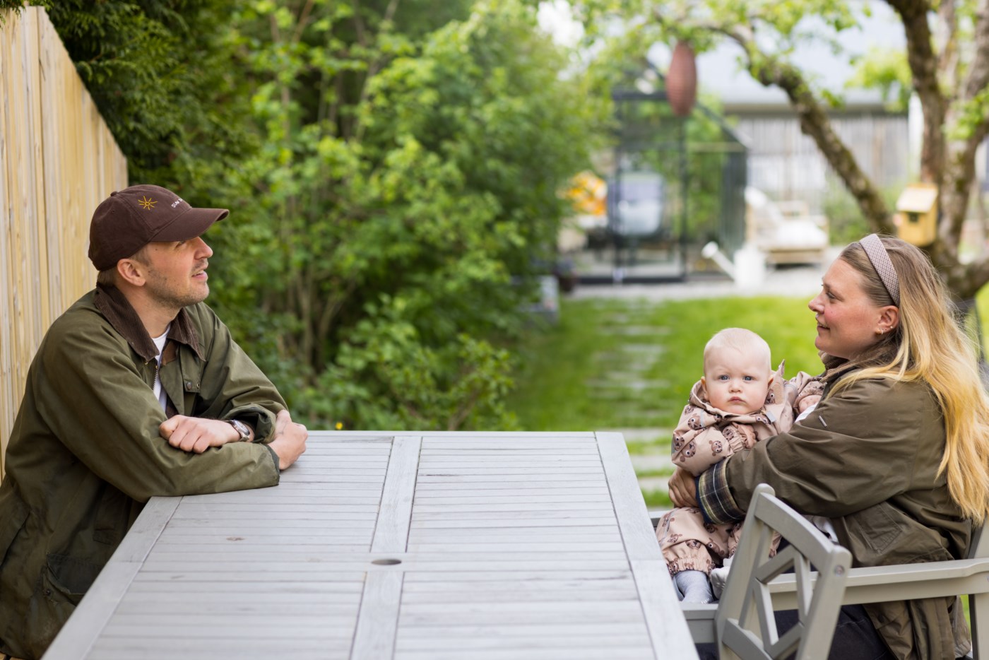 En mann med caps og en kvinne sitter på hver sin side av et bord. Kvinnen holder en baby.